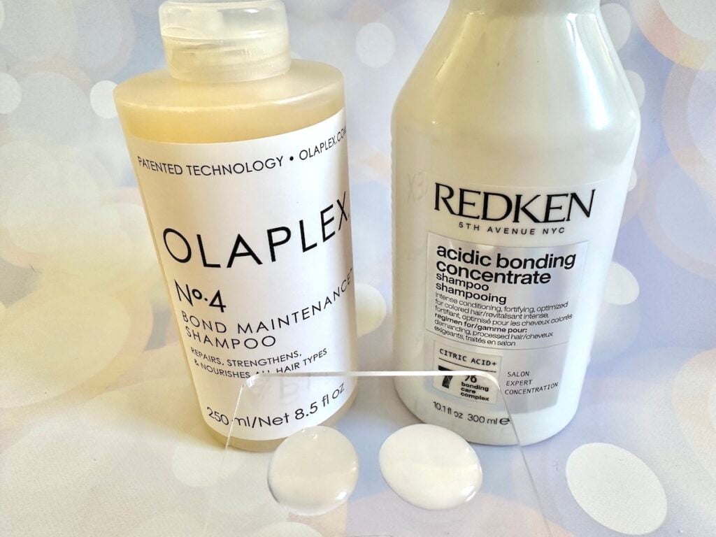 בקבוקי שמפו של Redken Acidic Bonding Concentrate ו-Olaplex No. 4 Bond Maintenance שמפו מאחורי דגימות על מרית פלסטיק שקופה.
